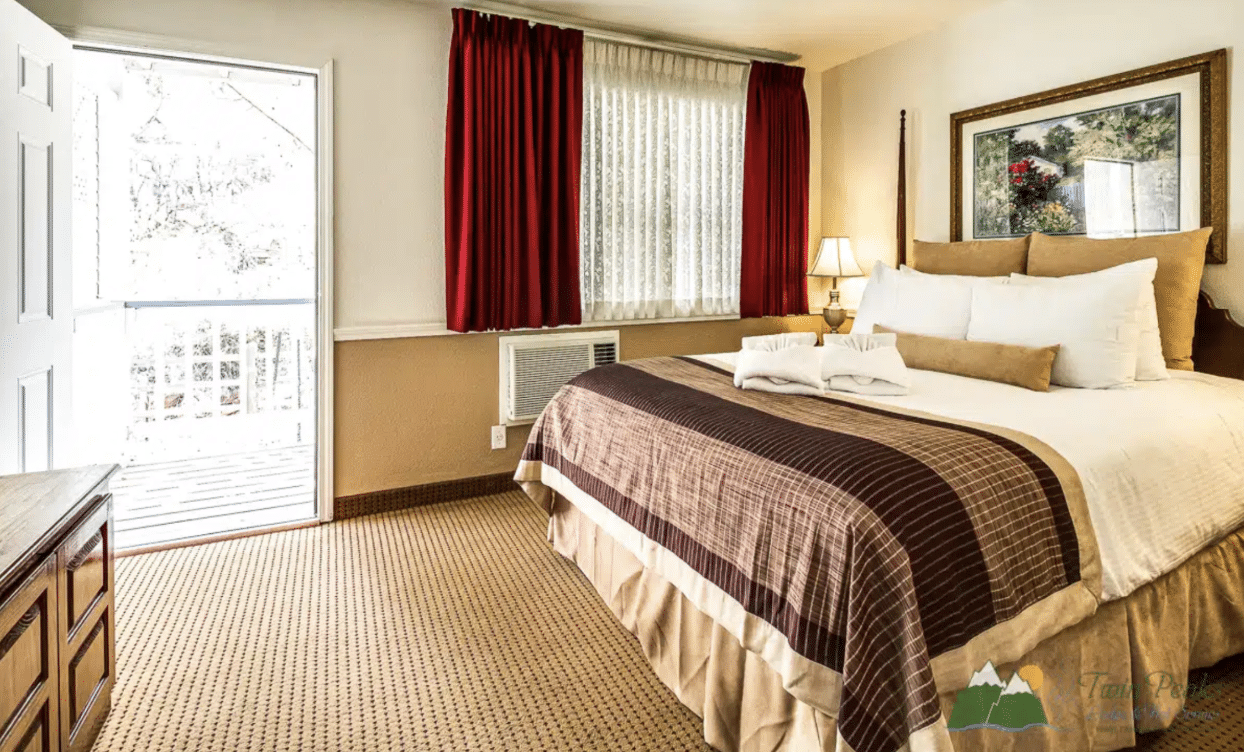 Spacious hotel bedroom with a king-size bed, white linens, burgundy curtains, bedside lamp, framed artwork, and a bright window overlooking the outdoors.